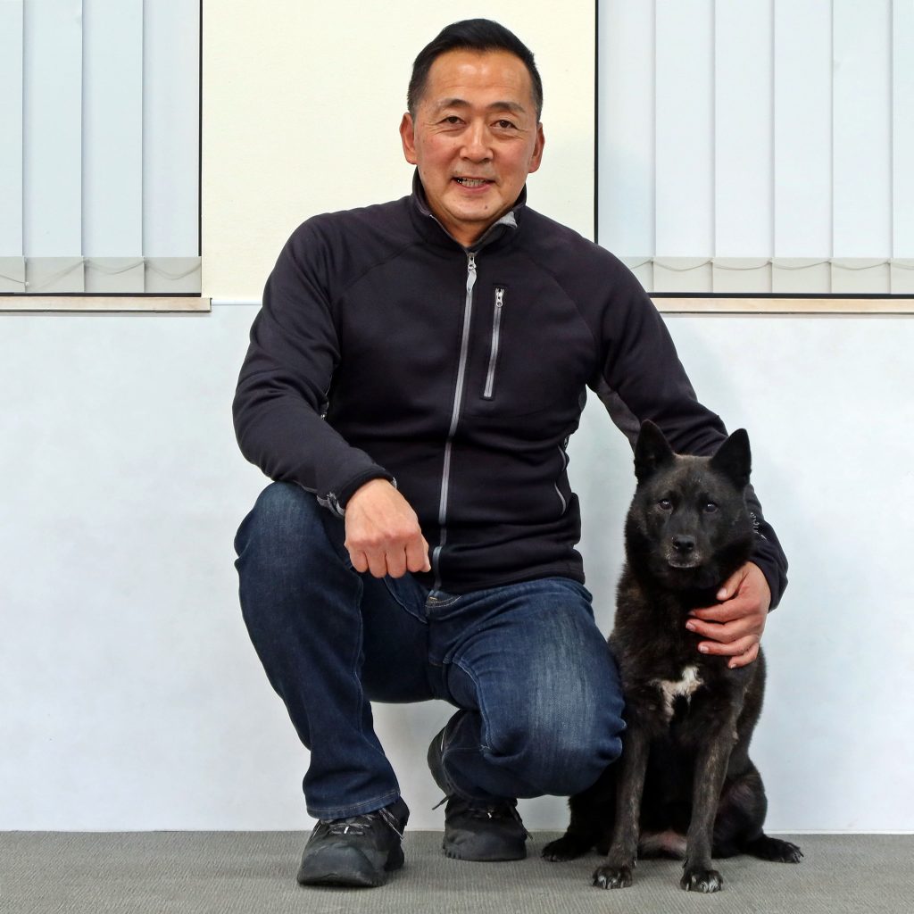 みなとみらいペットウェルネスクリニックで犬のしつけ教室を行うトレーナーが、黒い愛犬を横に笑顔しゃがんでいる。